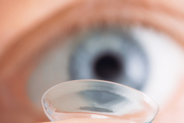A close up image of a person's eye with the eyecolour blue and a contact lens on their finger tip.