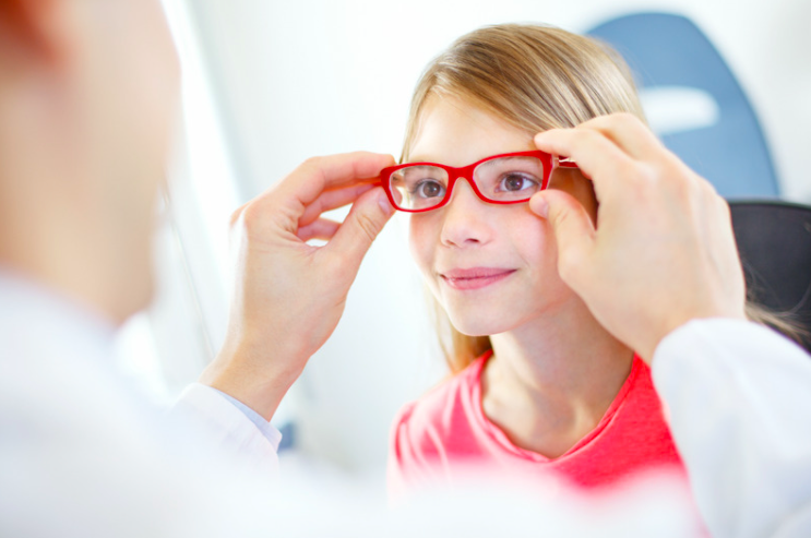 A young girl tries on her new red glasses at her eye exam